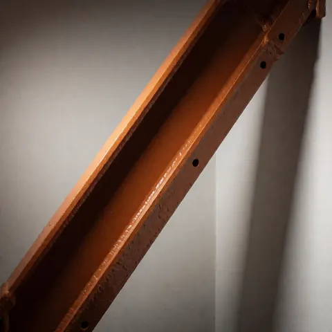 Boulon industriel en acier peint en orange, présentant des trous de vis et des soudures apparentes, posé contre un mur gris.