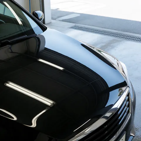 Capot d'une voiture noire polie reflétant des lumières fluorescentes dans une zone de lavage avec sol en béton et grille de drainage.