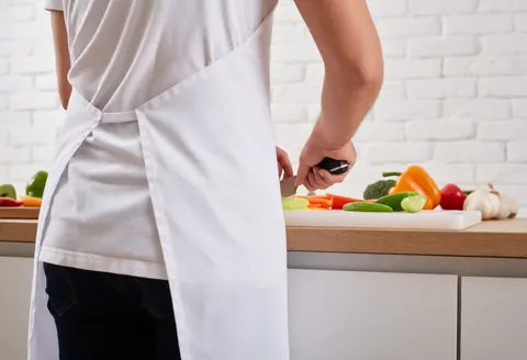 Une personne portant un tablier blanc coupe des légumes dans une cuisine moderne avec un mur en briques blanches visibles en arrière-plan.