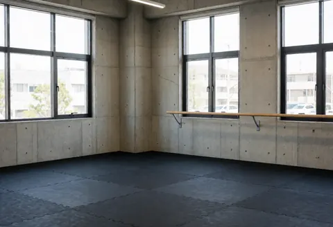 Studio de danse moderne avec murs en béton, grandes fenêtres et sol en tapis noirs, idéal pour les cours de danse.