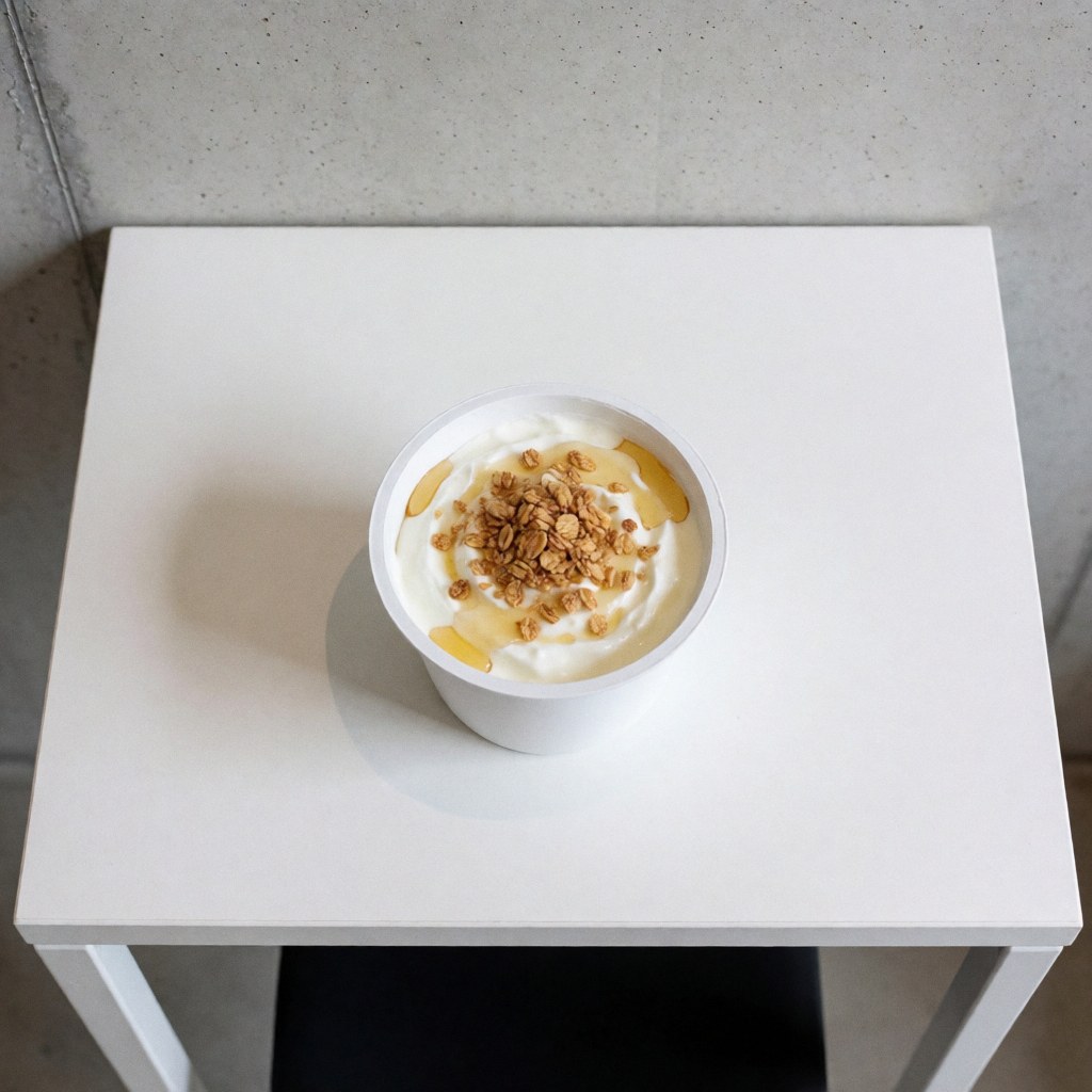 Yaourt grec dans une tasse blanche posée sur une table blanche, décoré de miel et de granola, dans un apart-hotel avec un mur en béton, vue de dessus.