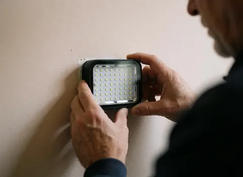 Gros plan sur les mains d'un technicien qui installe un projecteur LED avec des diodes visibles sur un mur beige, détail technique de l'équipement d'éclairage.