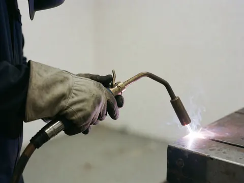 Une main portant un gant de cuir tient une torche de soudage à arc électrique en train de souder du métal dans un atelier industriel.
