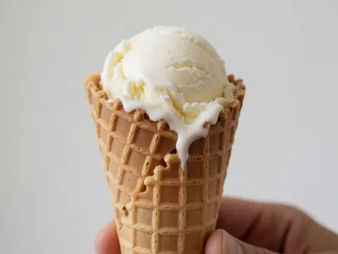 Glace vanille fondue tenue par une main dans un cône de gaufre sur fond blanc, idéal pour une pâtisserie ou un hôtel de luxe.