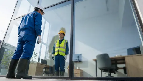 Un ouvrier en équipement de sécurité nettoie une grande vitre avec un reflet de bureau visible, illustrant un service de nettoyage professionnel en cours.