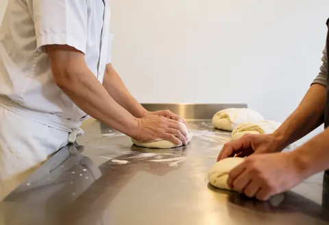 Un boulanger en uniforme blanc pétrissant la pâte sur un comptoir en acier inoxydable, avec de la farine visible et des pains à proximité dans une boulangerie propre.