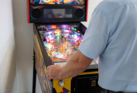 Un adulte joue au flipper dans un centre de loisirs, les mains sur les boutons, avec une piste illuminée par des lumières colorées.
