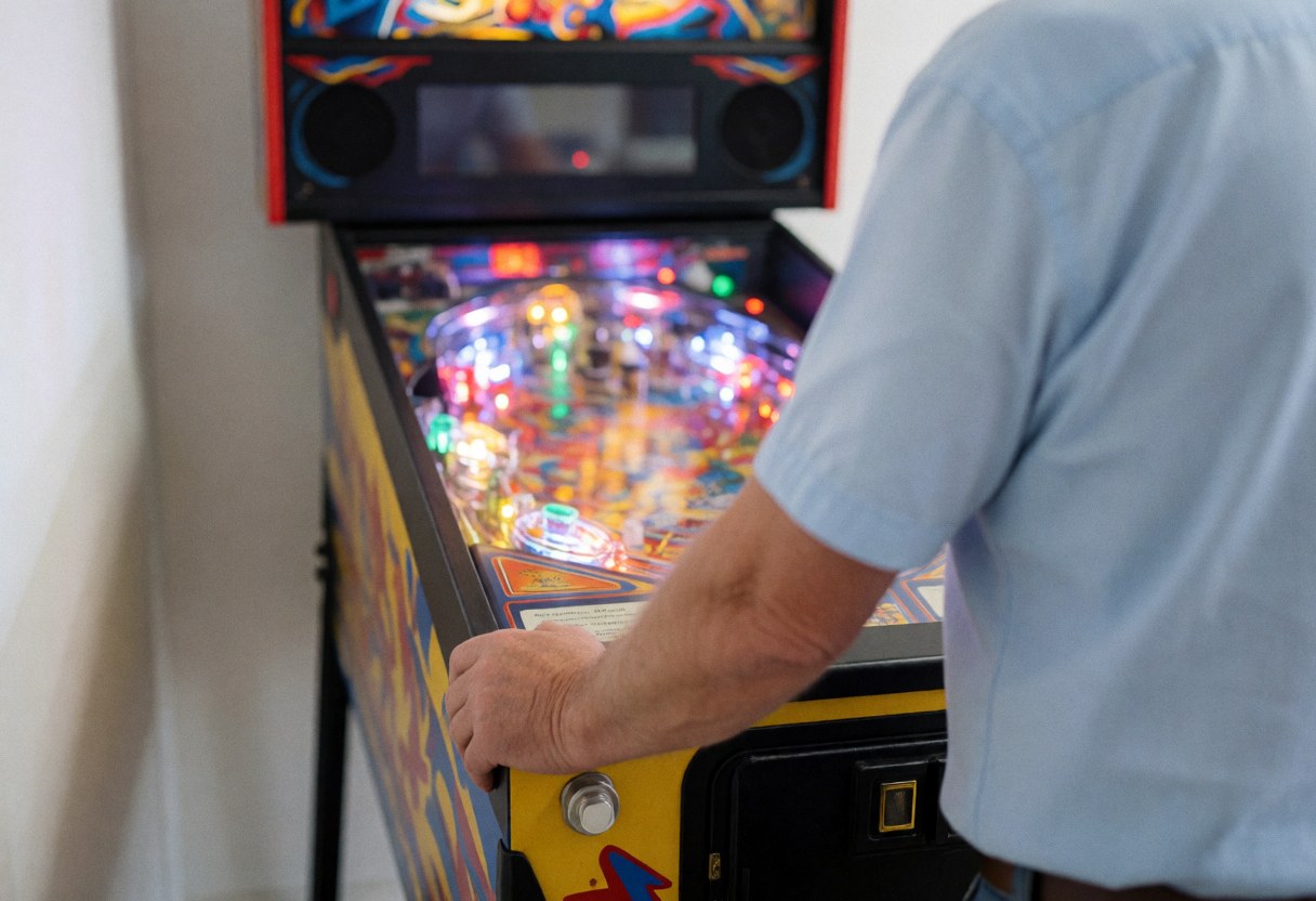 Un adulte joue au flipper dans un centre de loisirs, les mains sur les boutons, avec une piste illuminée par des lumières colorées.