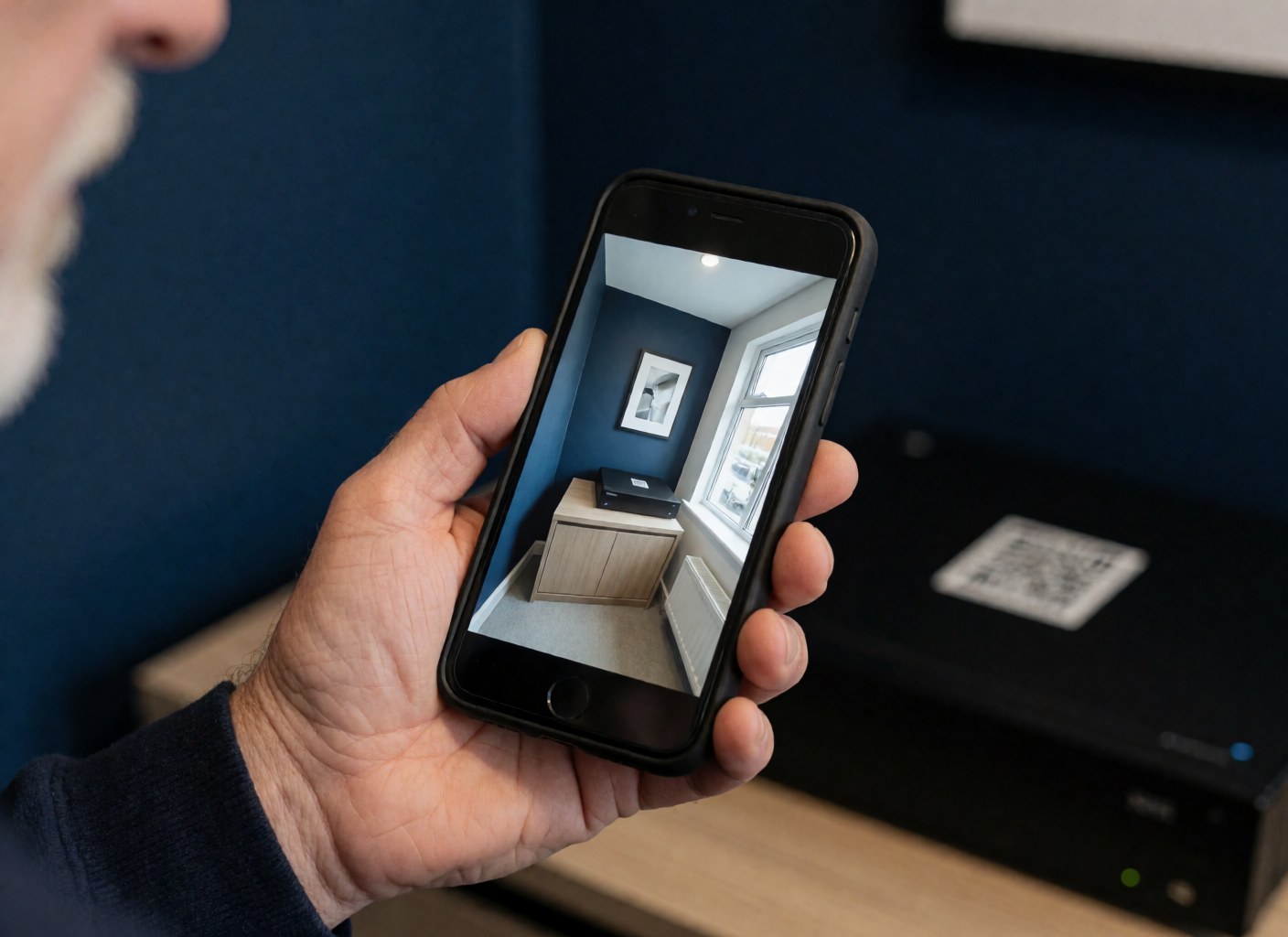 Une main tient un smartphone montrant l'image d'une caméra de surveillance dans une pièce aux murs bleu marine, avec un enregistreur vidéo numérique et un code QR visibles.