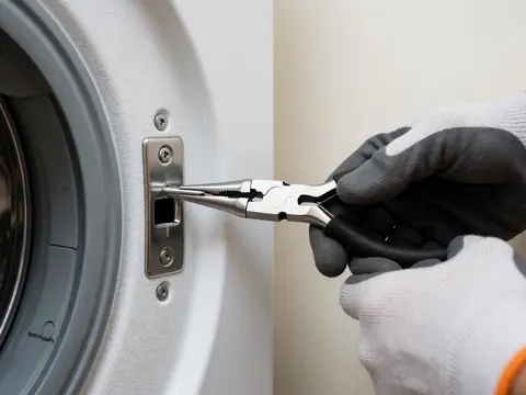 Gros plan d'une main portant un gant insérant un outil métallique dans la porte d'un lave-linge blanc, révélant l'intérieur texturé de l'appareil.