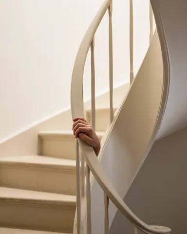 Une main repose sur la rampe en bois d'un escalier en colimaçon, devant un mur blanc et une lumière chaude, montrant un détail d'installation par un professionnel.