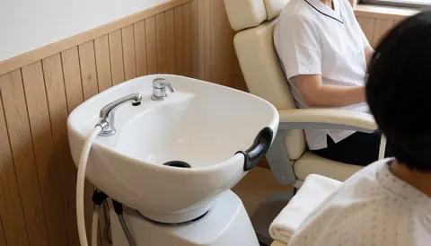 Un professionnel lave les cheveux d'un client dans un salon moderne. On voit un bac de lavage blanc, une chaise inclinable et un mur en bois clair dans un environnement propre et lumineux.