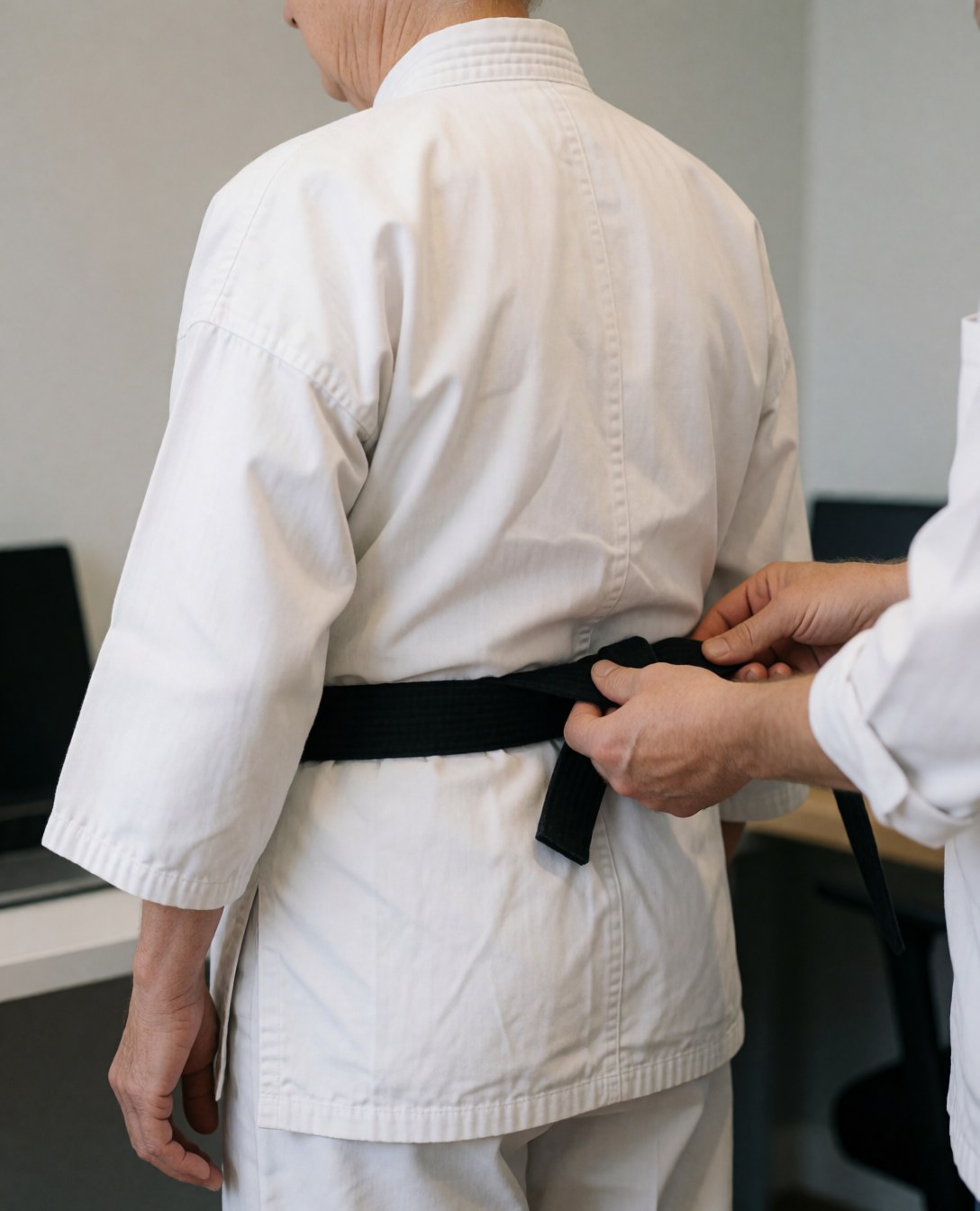 Instructeur de karate attachant une ceinture noire à un étudiant de dos dans un dojo aux murs gris clair.