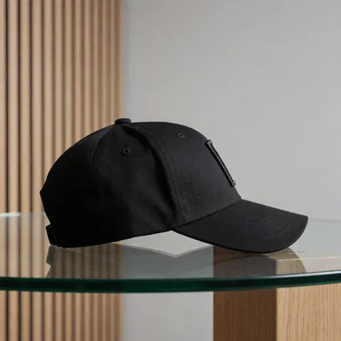 Casquette de baseball noire brodée posée sur une table en verre avec un fond gris et des panneaux en bois clair, idéale pour un catalogue de merchandising.