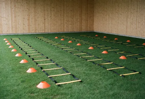Zone d'entraînement de football avec échelle d'agilité et cônes orange sur gazon synthétique, murs en bois, sans personnes.
