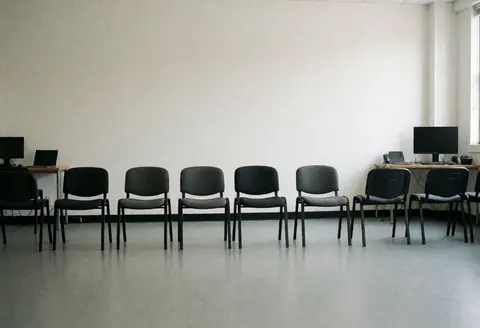 Chaises noires en rangée devant un mur blanc, tables avec ordinateurs, lumière naturelle, studio de danse prêt pour les auditions