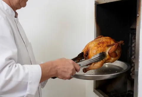 Un cuisinier retire un poulet rôti d'un four au charbon sur fond blanc, scène de service culinaire.