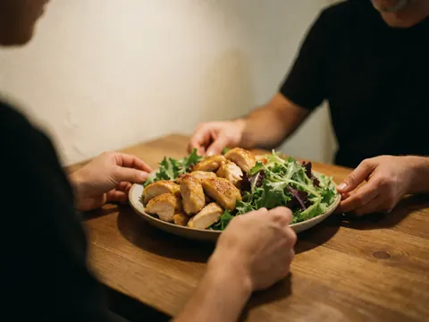 Des mains partagent un plat de poulet doré et de salade verte sur une table en bois dans un restaurant aux murs chaleureux.