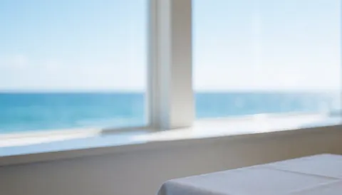 Table avec nappe blanche près d'une fenêtre offrant une vue sur la mer et des murs blancs, idéale pour un fond publicitaire de restaurant côtier