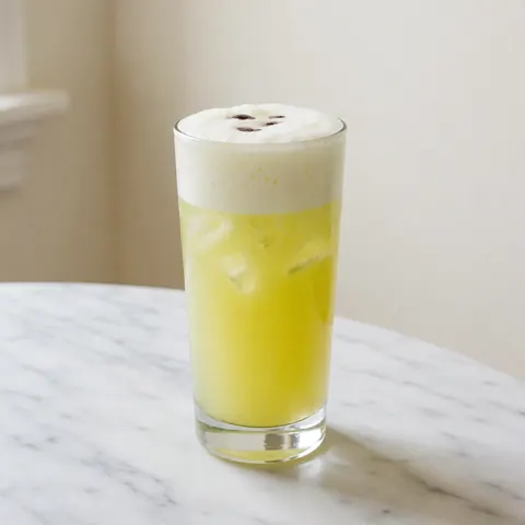 Un pisco sour servi dans un grand verre, garni de mousse blanche et de quelques gouttes d'amers, posé sur une table en marbre.