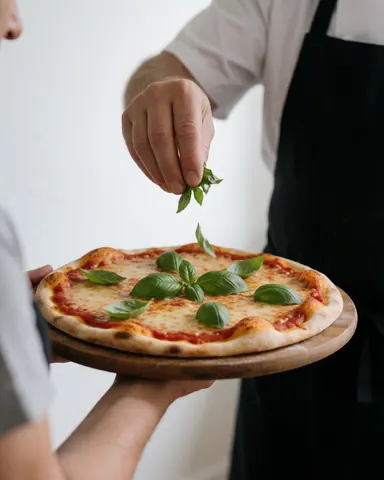 Le chef dépose des feuilles de basilic frais sur une pizza garnie de sauce et de fromage, posée sur une planche en bois devant un mur blanc.