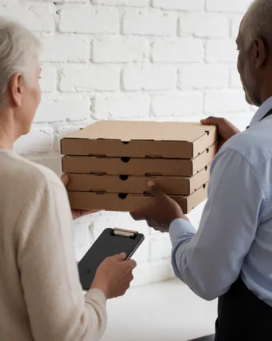 Un employé de pizzeria remet une pile de caisses de pizza à un client tenant un porte-documents dans une zone de retrait avec un mur en briques blanches.