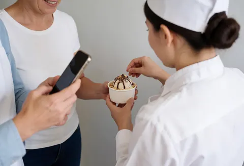 Un serveur ajuste des toppings au chocolat et aux noix sur une glace pendant qu'un client prend une photo dans une glacerie.