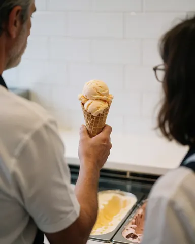 Une cliente reçoit une glace dans un cône en gaufre au comptoir d'une glacerie aux murs carrelés blancs.