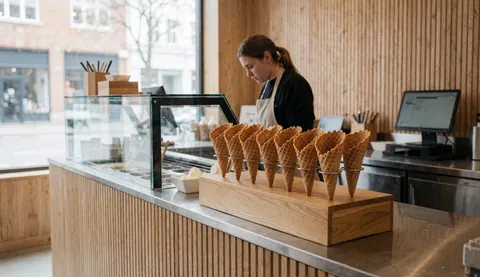 Des cônes de gaufre disposés sur le comptoir d'une crèmerie avec une personne travaillant en arrière-plan, des murs en bois et une caisse enregistreuse.